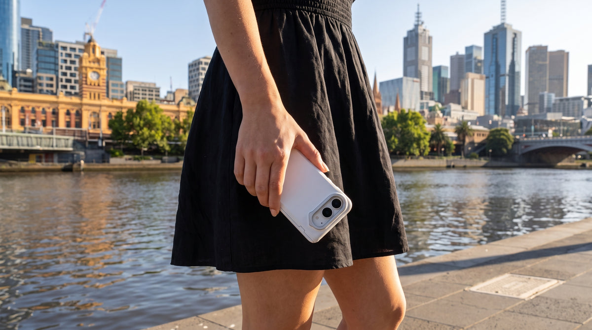 Woman in a black summer dress holding a white iPhone case, Melbourne waterfront skyline behind her